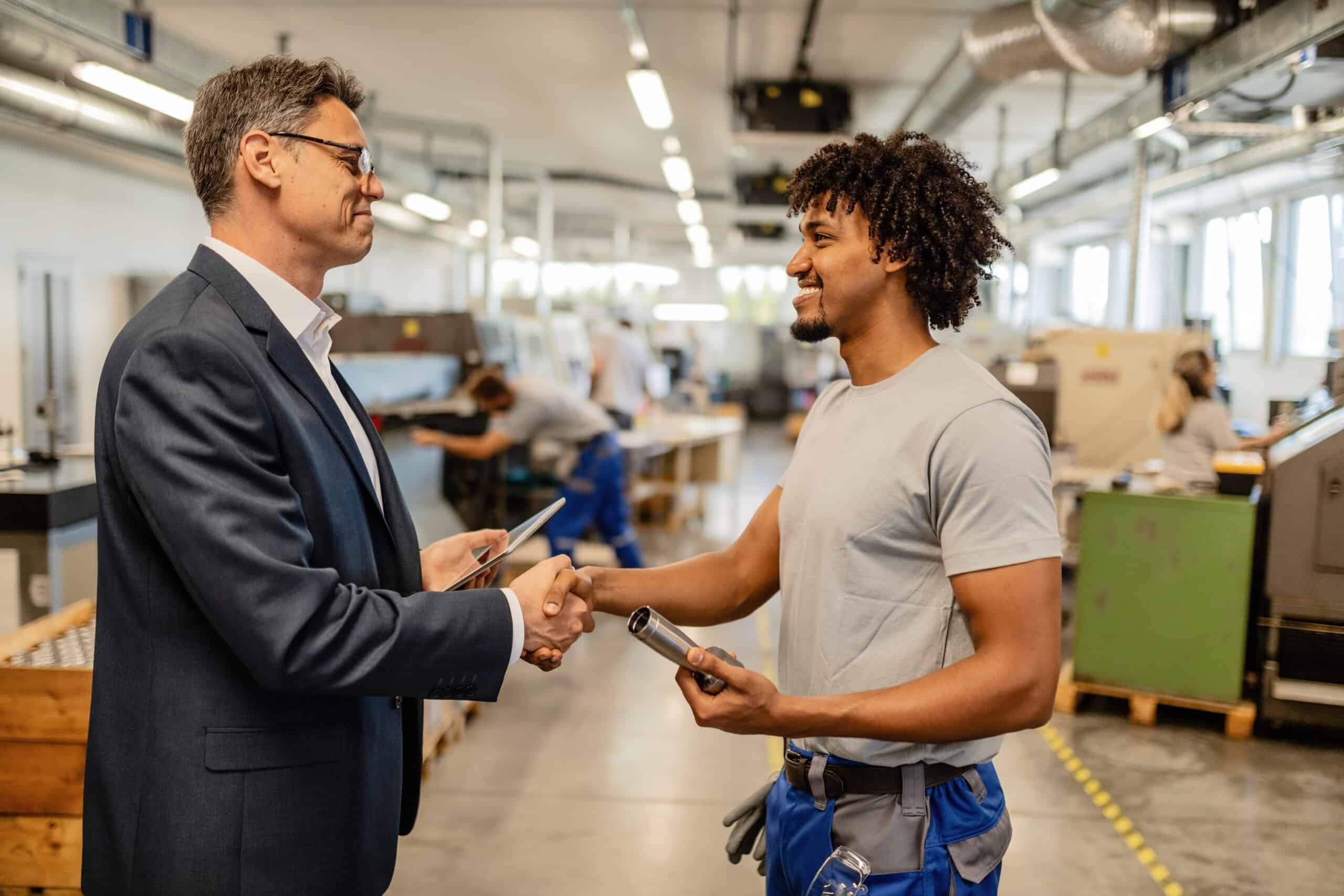 Happy businessman and African American manual worker shaking hands in factory plant.