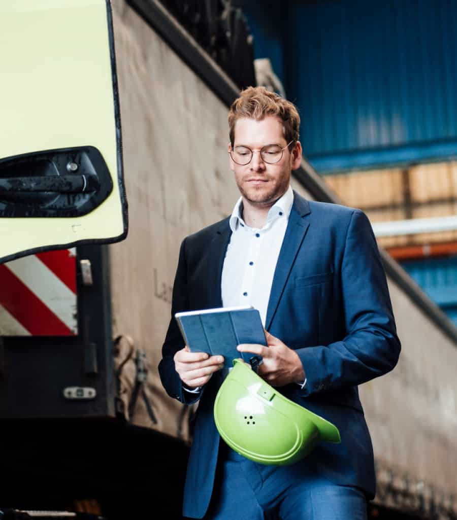 Man with glasses, he is wearing a suit and in his hands he has a tablet and a safety helmet.